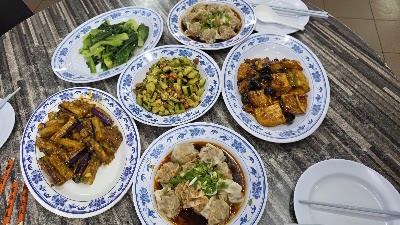San Gu Cuisine table spread featuring steamed dumplings, braised tofu with black olives, eggplant with garlic sauce, diced zucchini with corn, minced pork with mushrooms, and sautéed Chinese broccoli on traditional blue and white porcelain plates. — Items: Sautéed Chinese Broccoli · Minced Pork with Shiitake Mushrooms · Diced Zucchini with Corn and Mixed Vegetables · Braised Tofu with Black Olives · Eggplant with Garlic Sauce · Steamed Dumplings in Broth