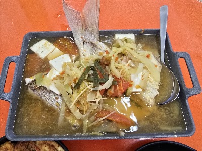 Fish head soup with tofu cubes, sliced vegetables, and whole fish in broth served in a black tray at Mid's Bakery restaurant. — Items: Fish Head Soup with Tofu and Vegetables · Whole Fish