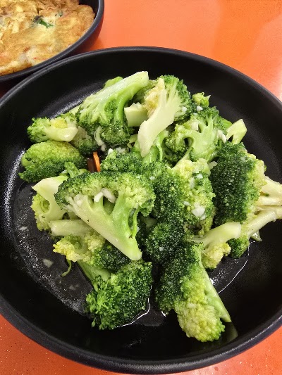 Stir-fried broccoli with garlic and sliced onions served in a black bowl at Mid's Bakery, with fried rice visible in the background. — Items: Broccoli with Garlic · Egg Fried Rice or similar Asian dish