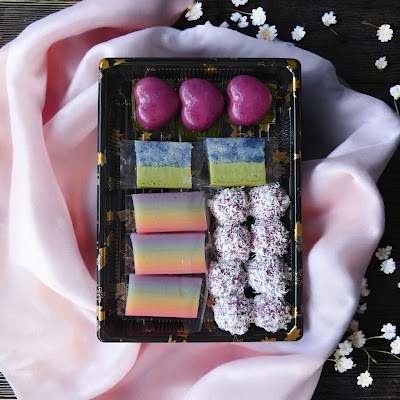 Kueharmony Nonya Kueh assorted pastries featuring purple macarons, ombre cake slices in blue-green and rainbow colors, and purple coconut rolls in black gift box. — Items: Purple Macarons · Blue and Green Ombre Cake Slice · Rainbow Ombre Cake Slices · Purple Coconut Rolls