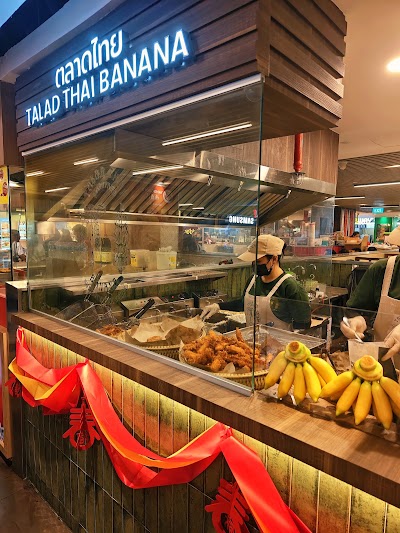 Talad Thai Banana food stall displaying fried chicken and fresh bananas at a Thai market counter with an employee preparing food. — Items: Fried Chicken · Bananas