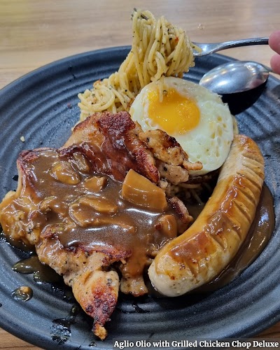 Aglio Olio with Grilled Chicken Chop Deluxe from Bradley's Grill features spaghetti, grilled chicken, sausage, fried egg, and caramelized onions on a dark plate. — Items: Aglio Olio with Grilled Chicken Chop Deluxe