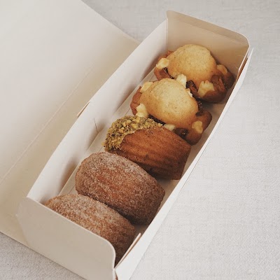 The Dessert Party assorted pastries box featuring cinnamon sugar cakes, white chocolate macadamia cookies, pistachio madeleine, chocolate madeleine, and cinnamon donut madeleine in white takeout packaging. — Items: Cinnamon Sugar Cake · White Chocolate Macadamia Nut Cookie · Pistachio Madeleine · Chocolate Madeleine · Cinnamon Donut Madeleine
