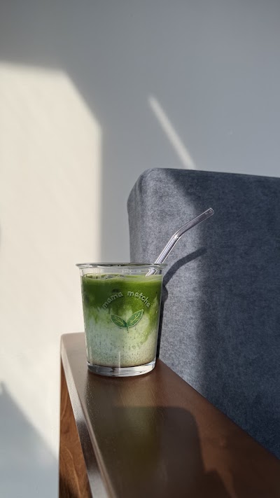 Mama Matcha iced matcha drink with glass straw in clear cup on wooden table with gray chair. — Items: Matcha Drink