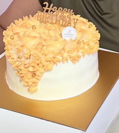 Happy Birthday cake with yellow buttercream rosettes and white fondant base, featuring 'Happy Birthday' gold topper and LadyLeFlour bakery label. — Items: Birthday Cake