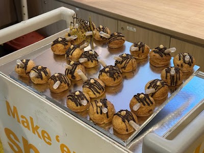 Display case of chocolate drizzle profiteroles with pearl decorations and a Happy Birthday cake topper from LadyLeFlour bakery. — Items: Chocolate Drizzle Profiterole · Happy Birthday Cake